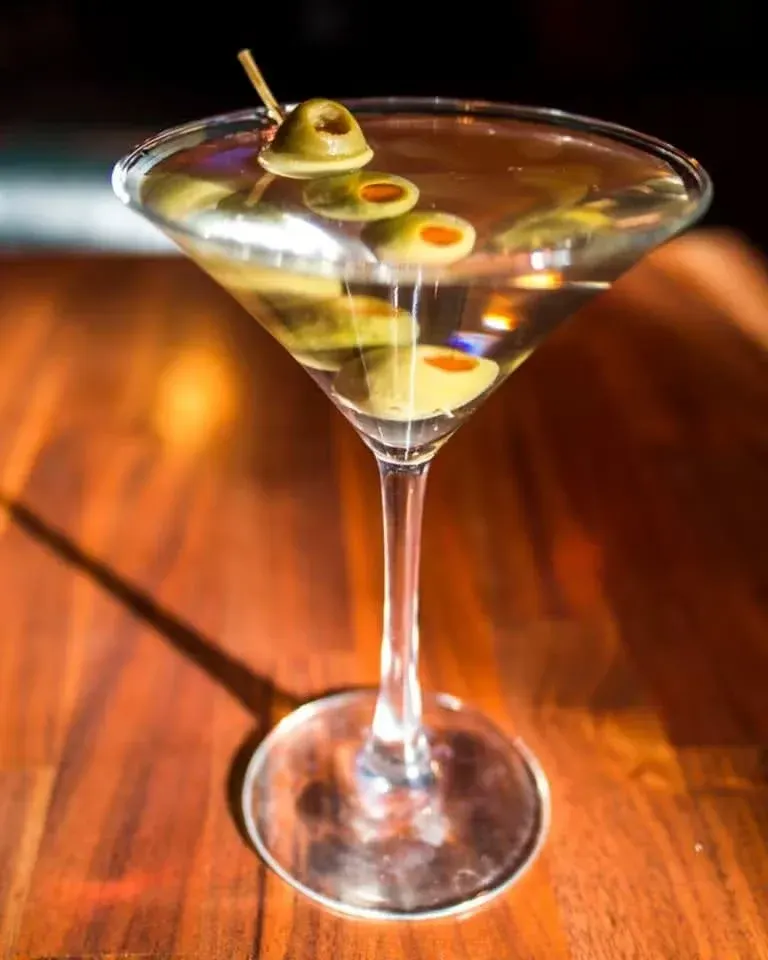A martini glass filled with clear liquid, garnished with green olives on a toothpick, on a wooden surface.