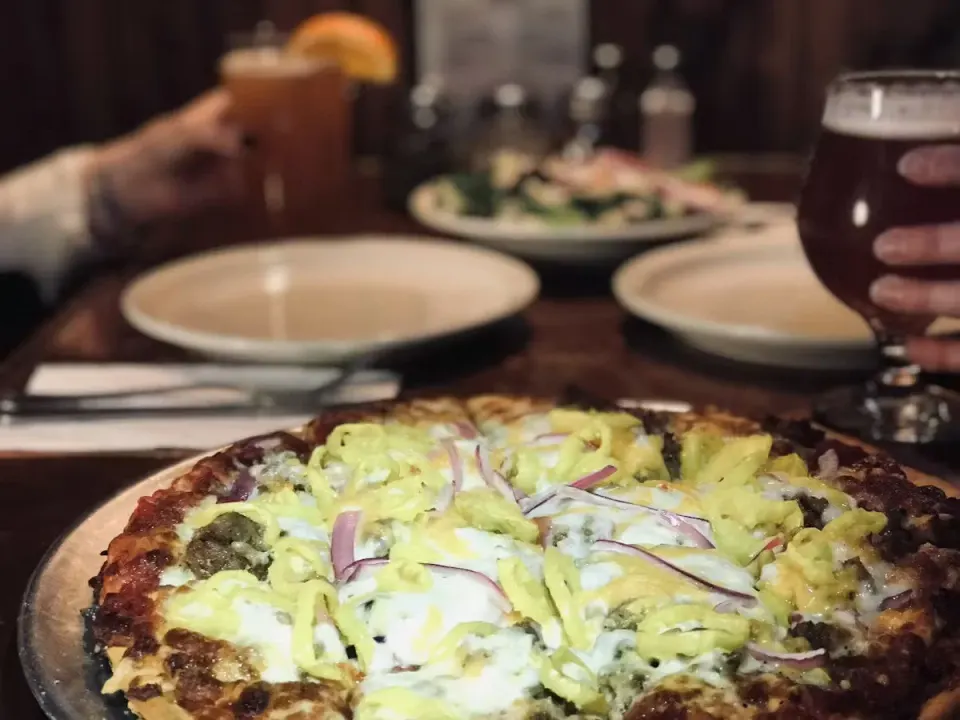 A pizza with toppings including green bell peppers, onions, and possibly sausage on a wooden table. In the background, people are holding drinks, including a glass of dark beer and a cocktail with an orange slice. There are empty plates and utensils on the table.