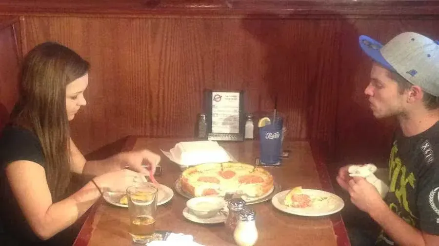 A woman and a man sitting at a restaurant table with a cheese pizza, glasses of water, and a blue drink with a lemon slice. The woman is cutting her food, and the man is holding a piece of bread.